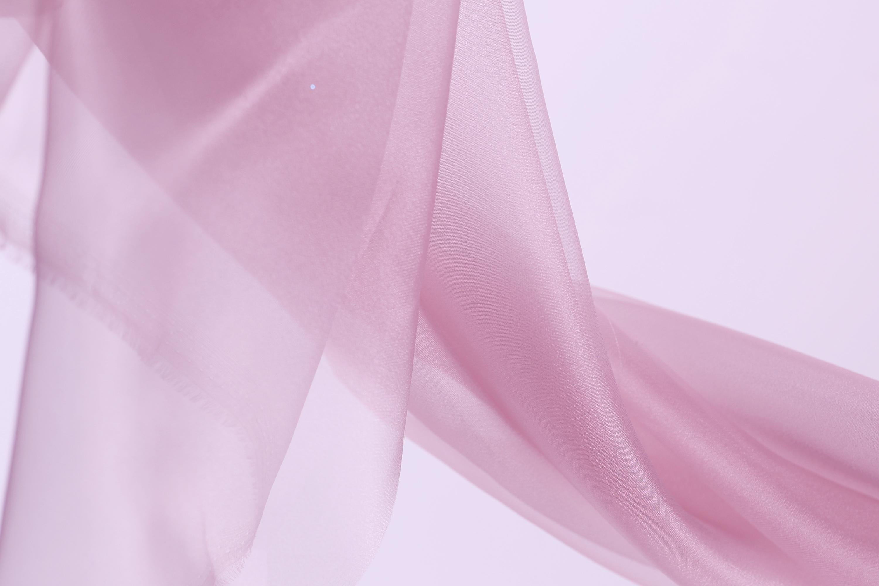 A close-up view of a soft, flowing pink fabric, possibly silk or chiffon, against a plain white background.
