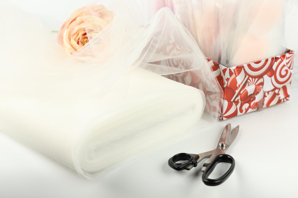 a white tulle fabric, a pair of scissors, and a pink rose, all arranged on a white surface.