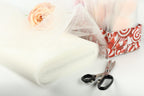 a white tulle fabric, a pair of scissors, and a pink rose, all arranged on a white surface.