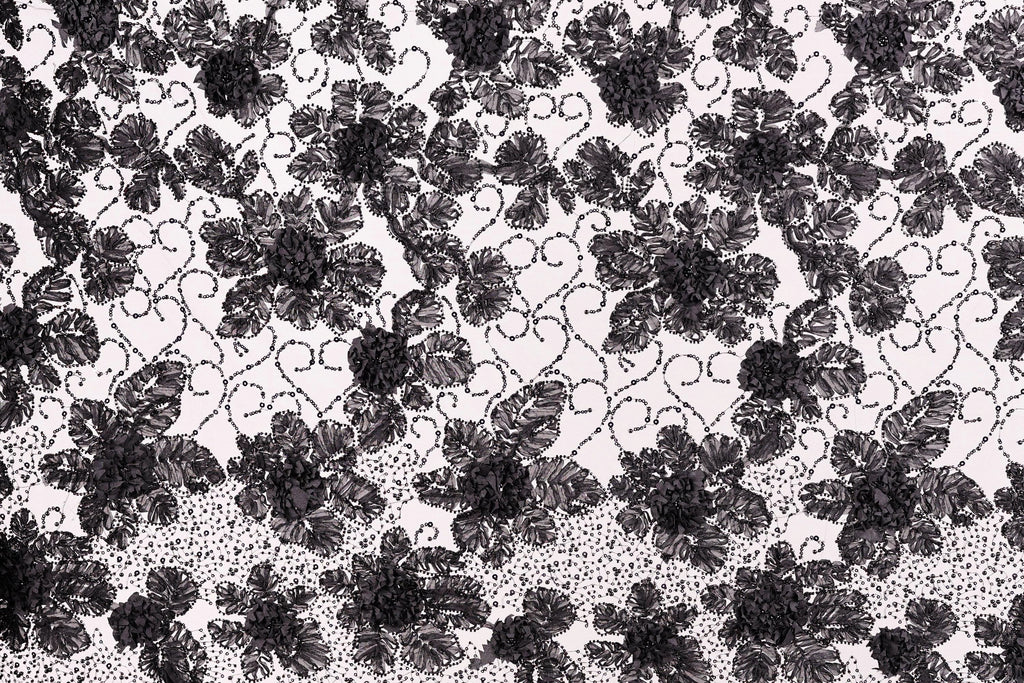 a close-up view of a black and white lace pattern with a repeating floral design. The lace appears to be made of a delicate material, possibly silk or chiffon, and is adorned with numerous small black flowers or leaves. The pattern is intricate and detailed, showcasing the craftsmanship and artistry involved in creating this type of lace.