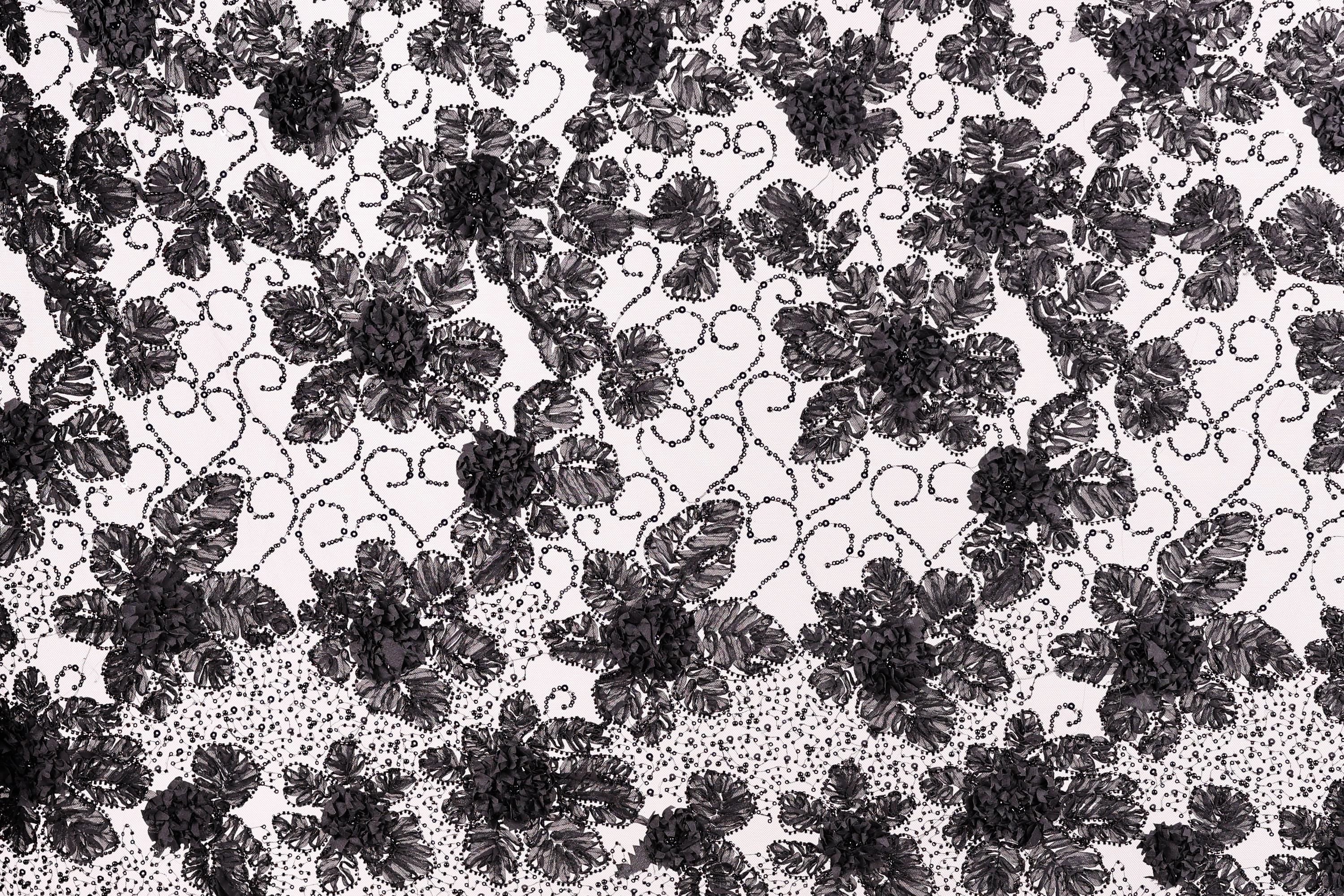 a close-up view of a black and white lace pattern with a repeating floral design. The lace appears to be made of a delicate material, possibly silk or chiffon, and is adorned with numerous small black flowers or leaves. The pattern is intricate and detailed, showcasing the craftsmanship and artistry involved in creating this type of lace.