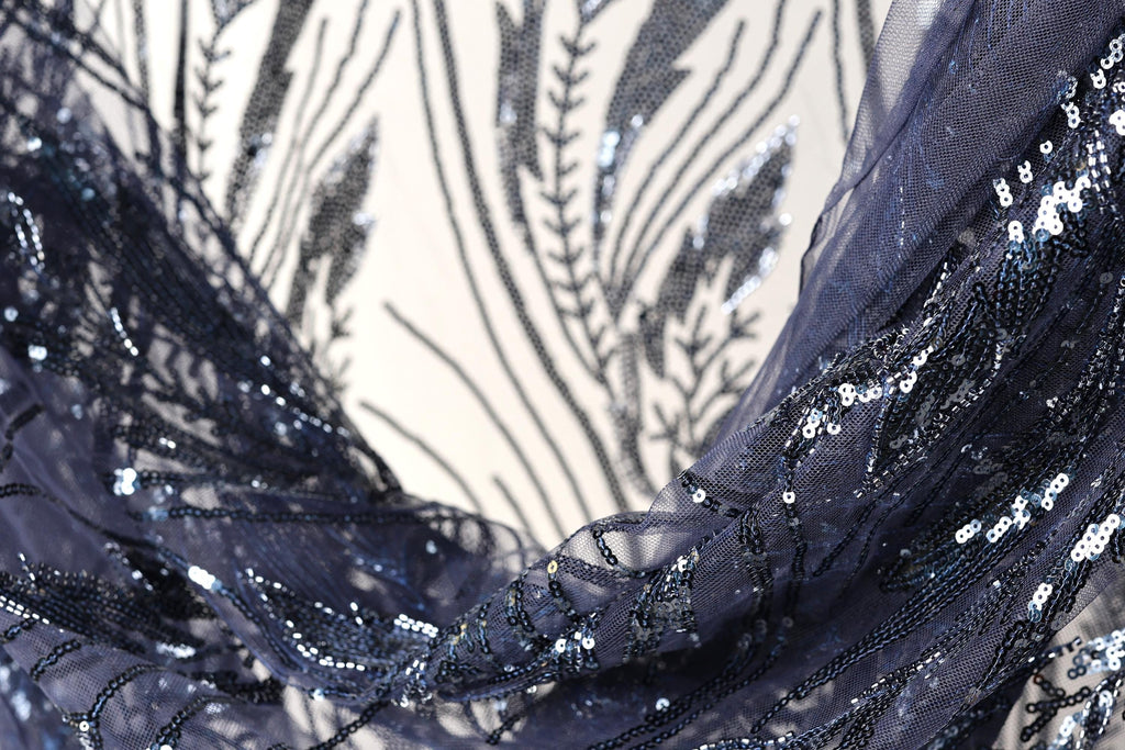 a close-up view of a fabric with a dark blue, sequined pattern. The fabric appears to be made of a sheer material, with a delicate and intricate design featuring small sequins and beads. The background is blurred, drawing focus to the fabric and its details.
