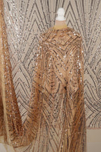 a close-up view of a sequin fabric with a gold and black pattern. The fabric appears to be a sheer material, with a metallic sheen and a textured appearance. The background is a plain white wall with a geometric pattern.