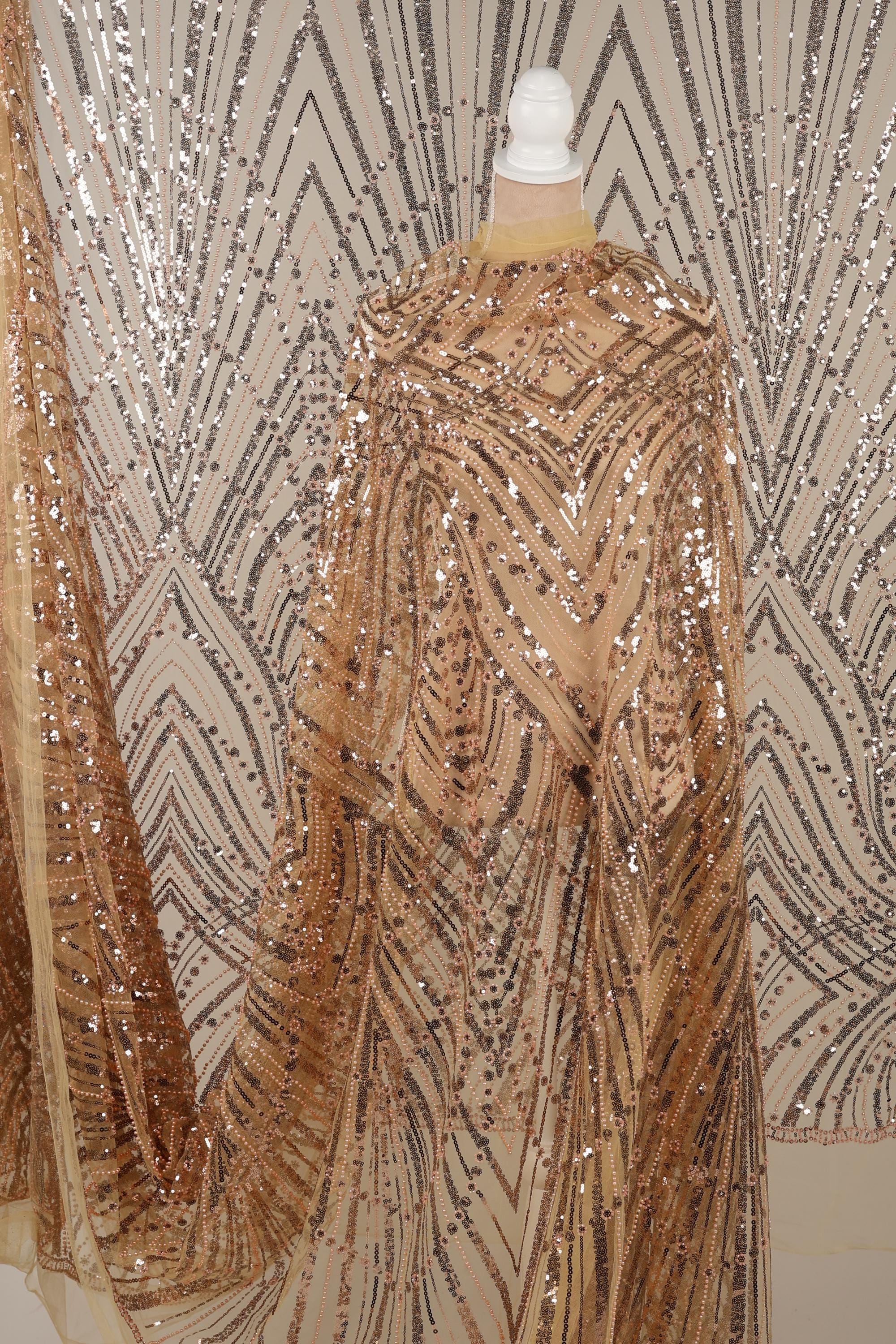 a close-up view of a sequin fabric with a gold and black pattern. The fabric appears to be a sheer material, with a metallic sheen and a textured appearance. The background is a plain white wall with a geometric pattern.