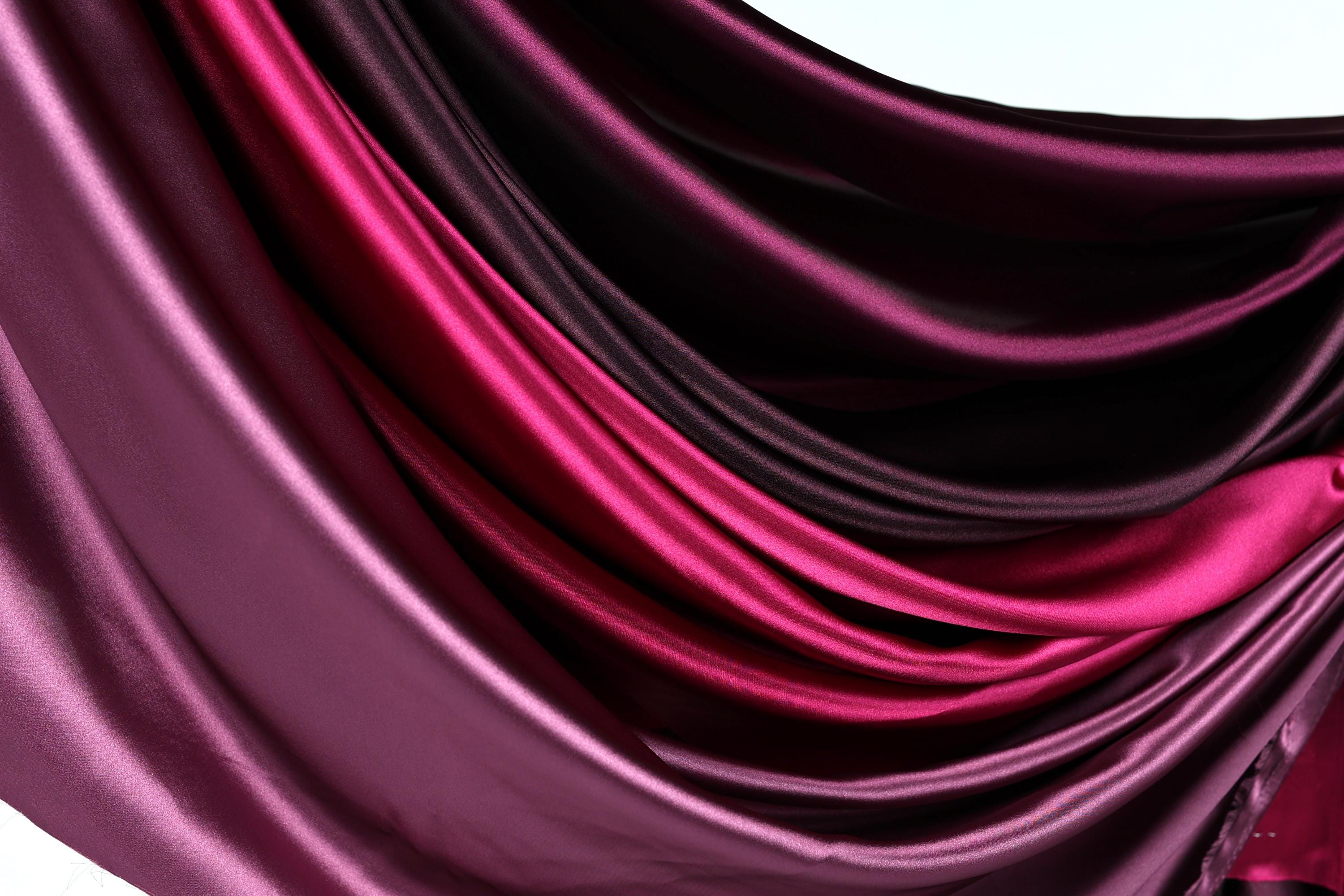 a close-up view of a fabric with a gradient of colors, transitioning from a deep purple to a lighter shade of purple. The fabric appears to be made of satin or a similar smooth, shiny material.