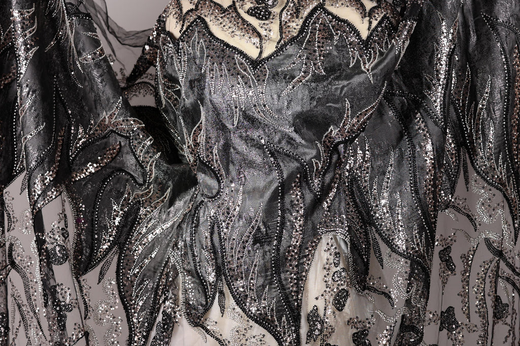 a close-up view of a black and silver fabric with intricate, sparkling designs. The fabric appears to be a sheer material, possibly lace or a similar type of textile. The design features a mix of black and silver elements, including sequins, beads, and floral patterns, creating a visually striking and detailed appearance.