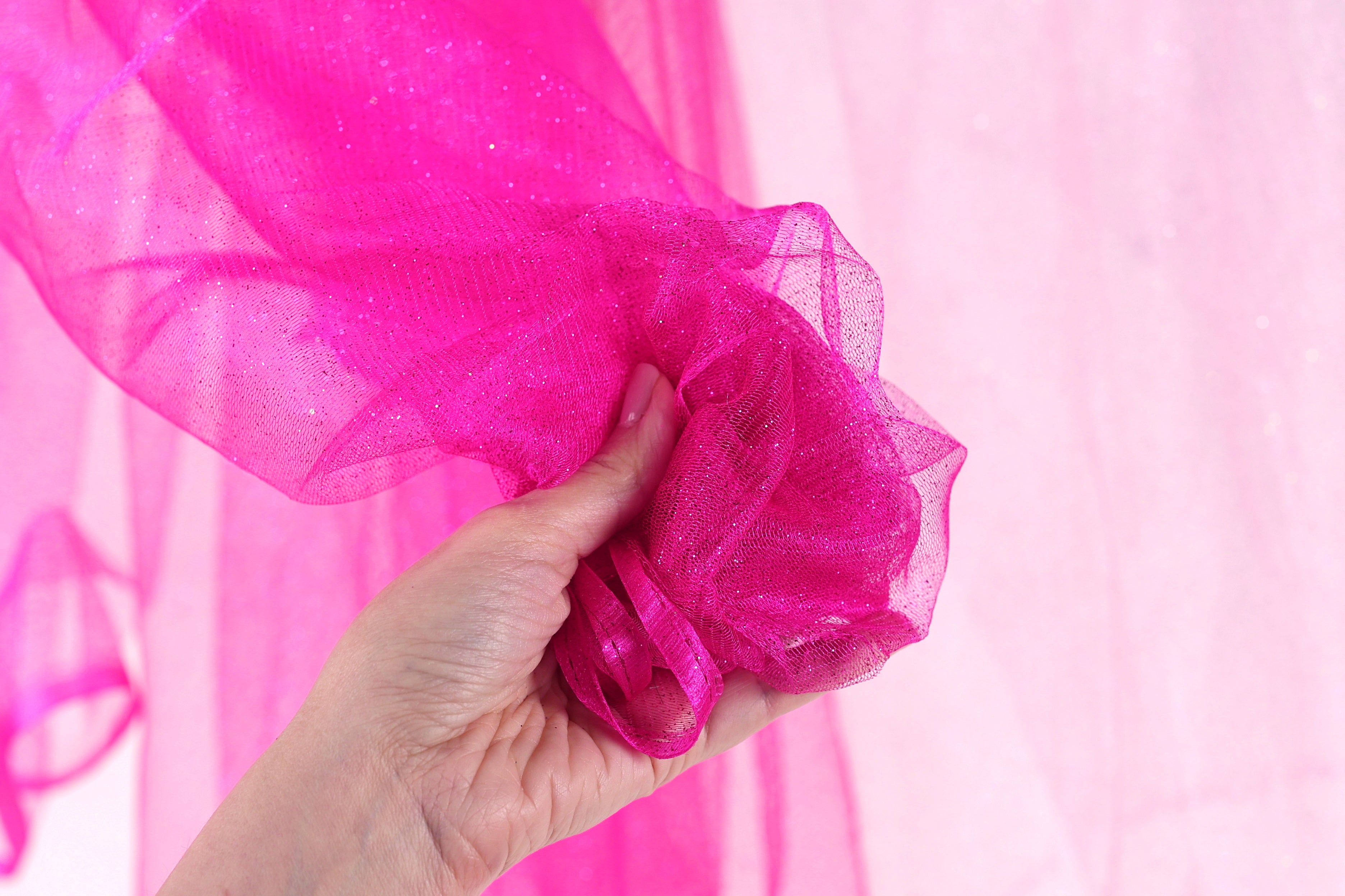 A hand is holding a pink, shiny fabric against a pink background.