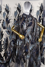 a mannequin wearing an elaborate, intricately designed cape or shawl with a floral pattern in shades of blue and gold. The cape has gold accents and appears to be made of a shiny, textured fabric.