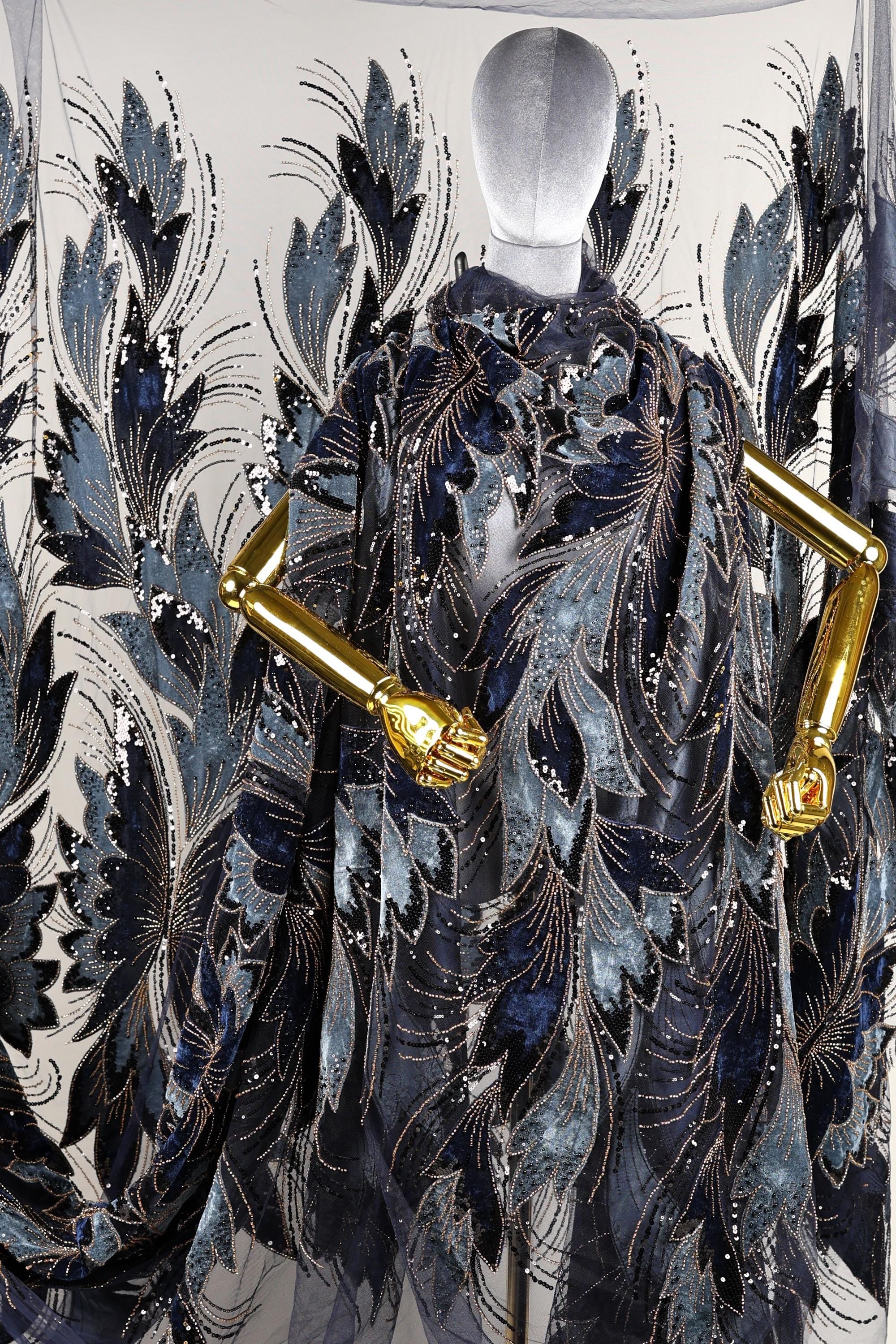 a mannequin wearing an elaborate, intricately designed cape or shawl with a floral pattern in shades of blue and gold. The cape has gold accents and appears to be made of a shiny, textured fabric.