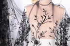 a mannequin wearing a dress with a sheer, black lace overlay. The lace has a floral pattern and is attached to the dress in a decorative manner.