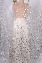 A mannequin wearing a white dress with silver leaf-like patterns on the skirt and a sheer white cape draped over the shoulders.