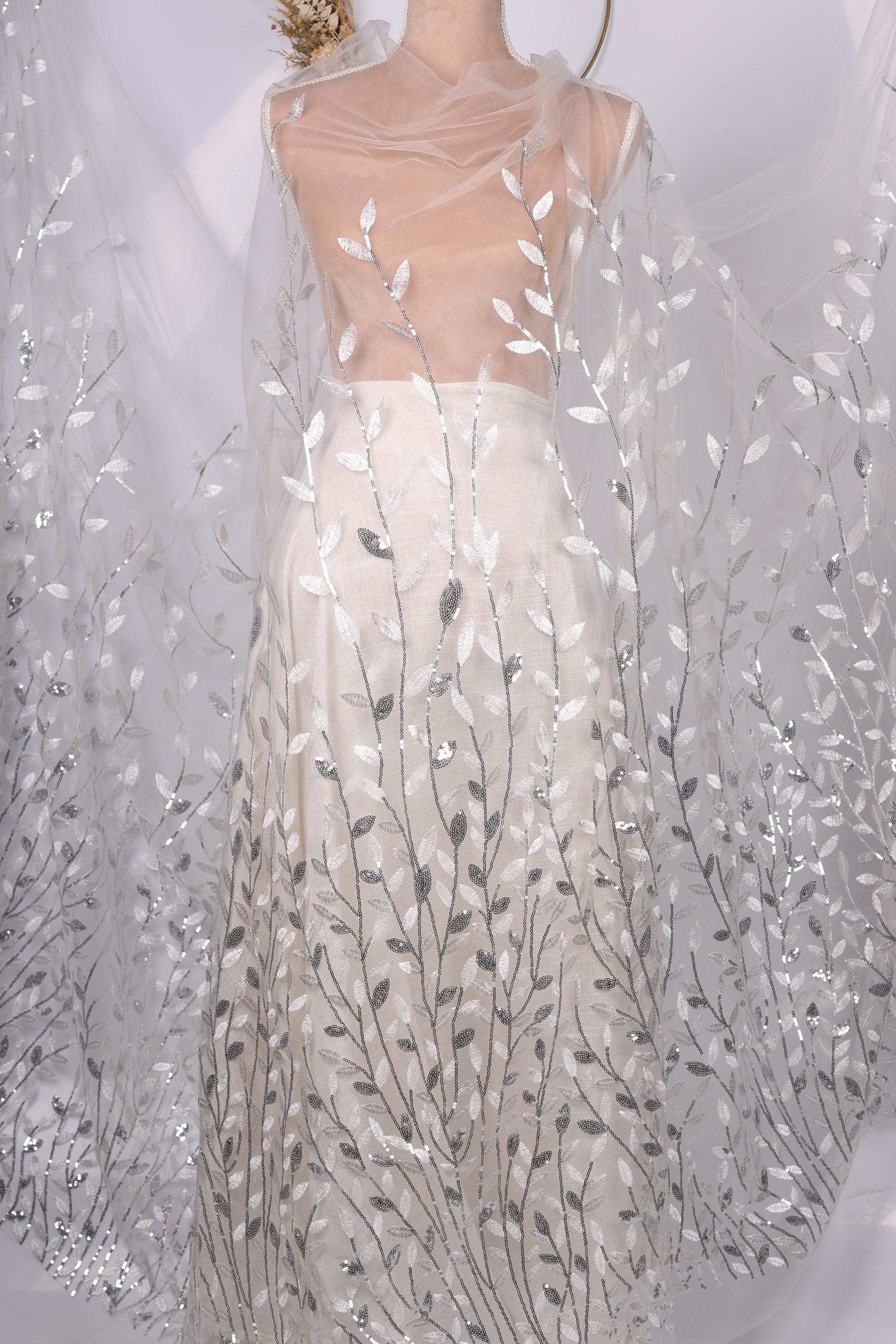 A mannequin wearing a white dress with silver leaf-like patterns on the skirt and a sheer white cape draped over the shoulders.