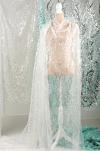 A mannequin wearing a white lace dress with a long, flowing cape draped over it, set against a backdrop of shimmering silver and turquoise sequins.