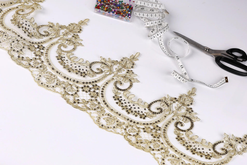 a close-up view of a decorative gold lace trim with intricate floral patterns, along with a pair of scissors and a measuring tape on a white background.