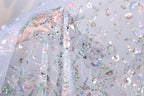 a close-up view of a delicate, intricately embroidered fabric with a floral pattern. The fabric appears to be made of sheer material, allowing a clear view of the detailed embroidery work. The colors used in the embroidery are predominantly pink, blue, and green, creating a visually appealing and vibrant design. The fabric is spread out, showcasing the full extent of the embroidered pattern.