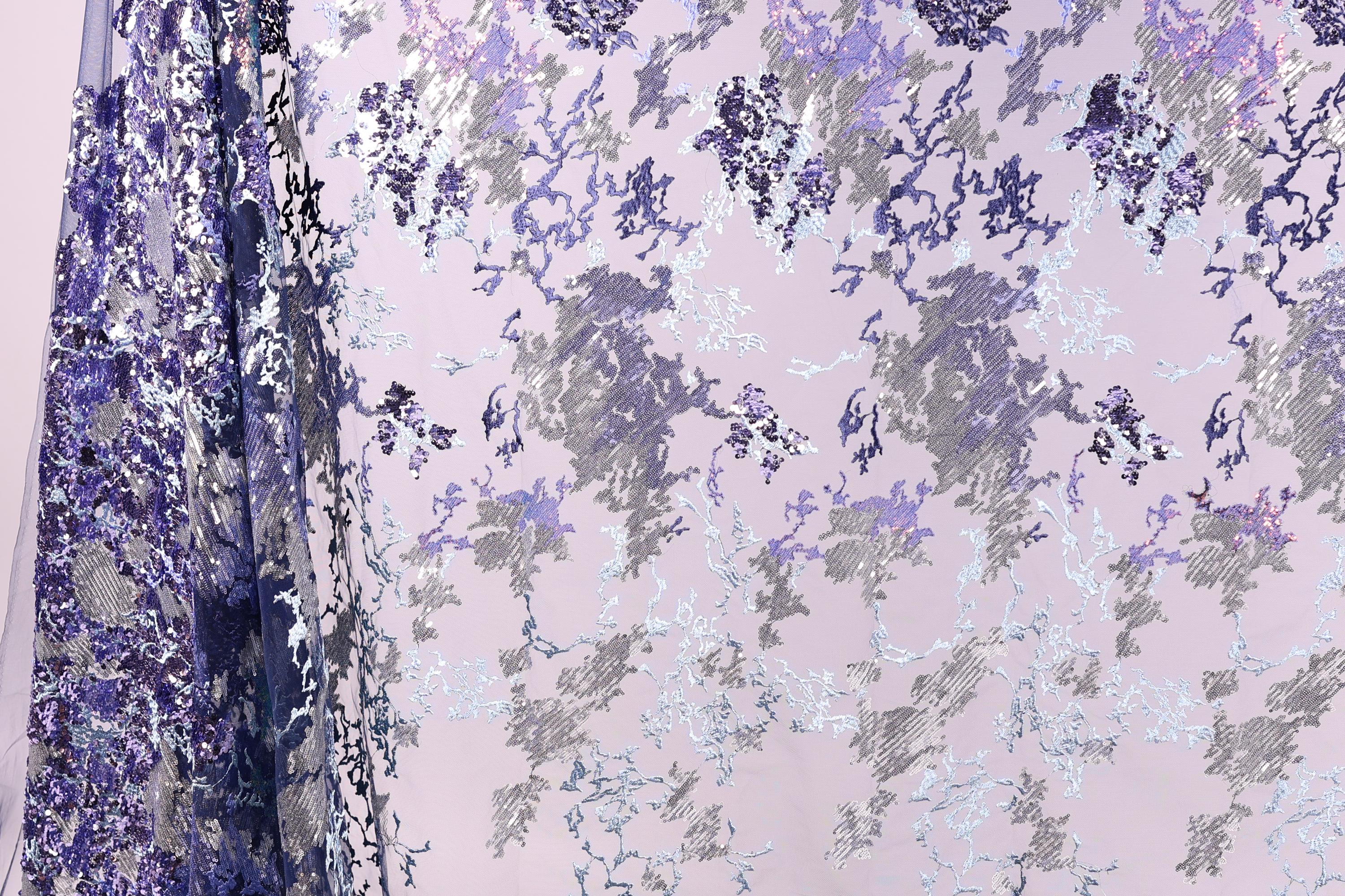 a fabric with a floral pattern in shades of purple and gray, with some white accents. The fabric appears to be a sheer or sheer-like material, with a delicate and intricate design.