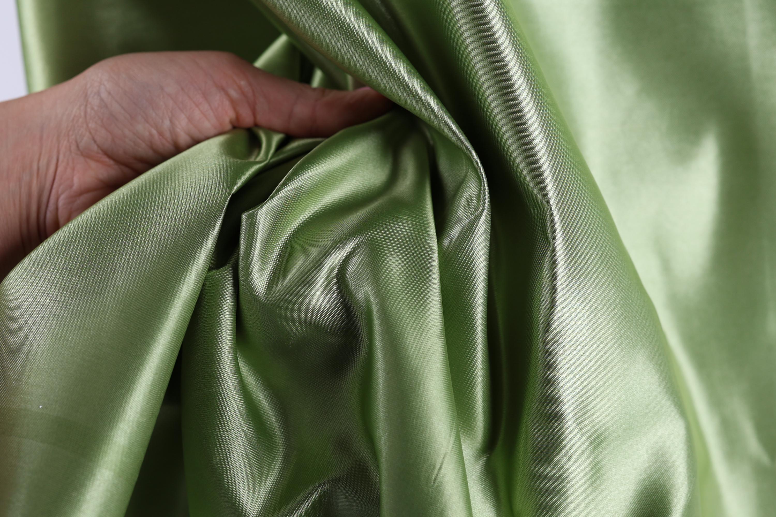 A hand is holding a piece of green satin fabric, which appears to be shiny and smooth.