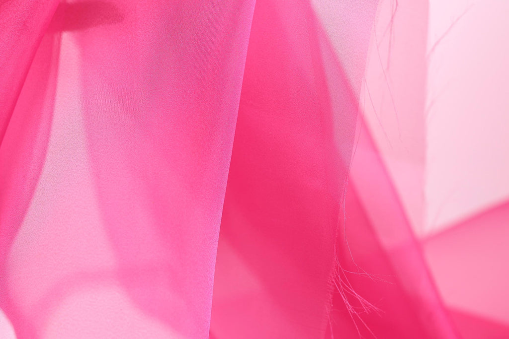 The image is a close-up of a soft, flowing fabric in a vibrant shade of pink. The fabric appears to be made of silk or chiffon, with a delicate and elegant texture. The background is blurred, drawing focus to the pink fabric and creating a visually striking contrast.