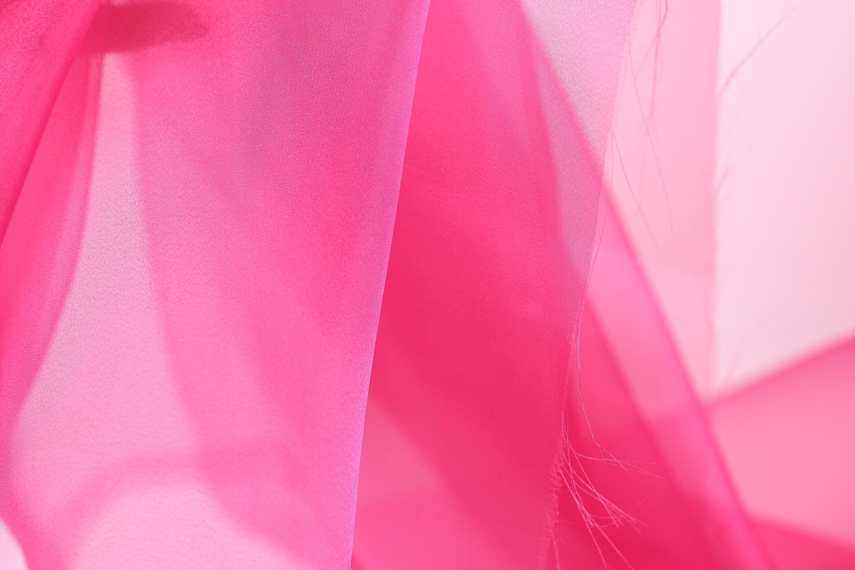 The image is a close-up of a soft, flowing fabric in a vibrant shade of pink. The fabric appears to be made of silk or chiffon, with a delicate and elegant texture. The background is blurred, drawing focus to the pink fabric and creating a visually striking contrast.