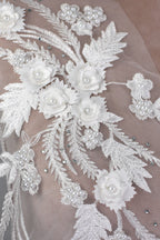 The image is a close-up view of a delicate, intricate white lace fabric adorned with numerous small white flowers and leaves, as well as silver rhinestones or beads.