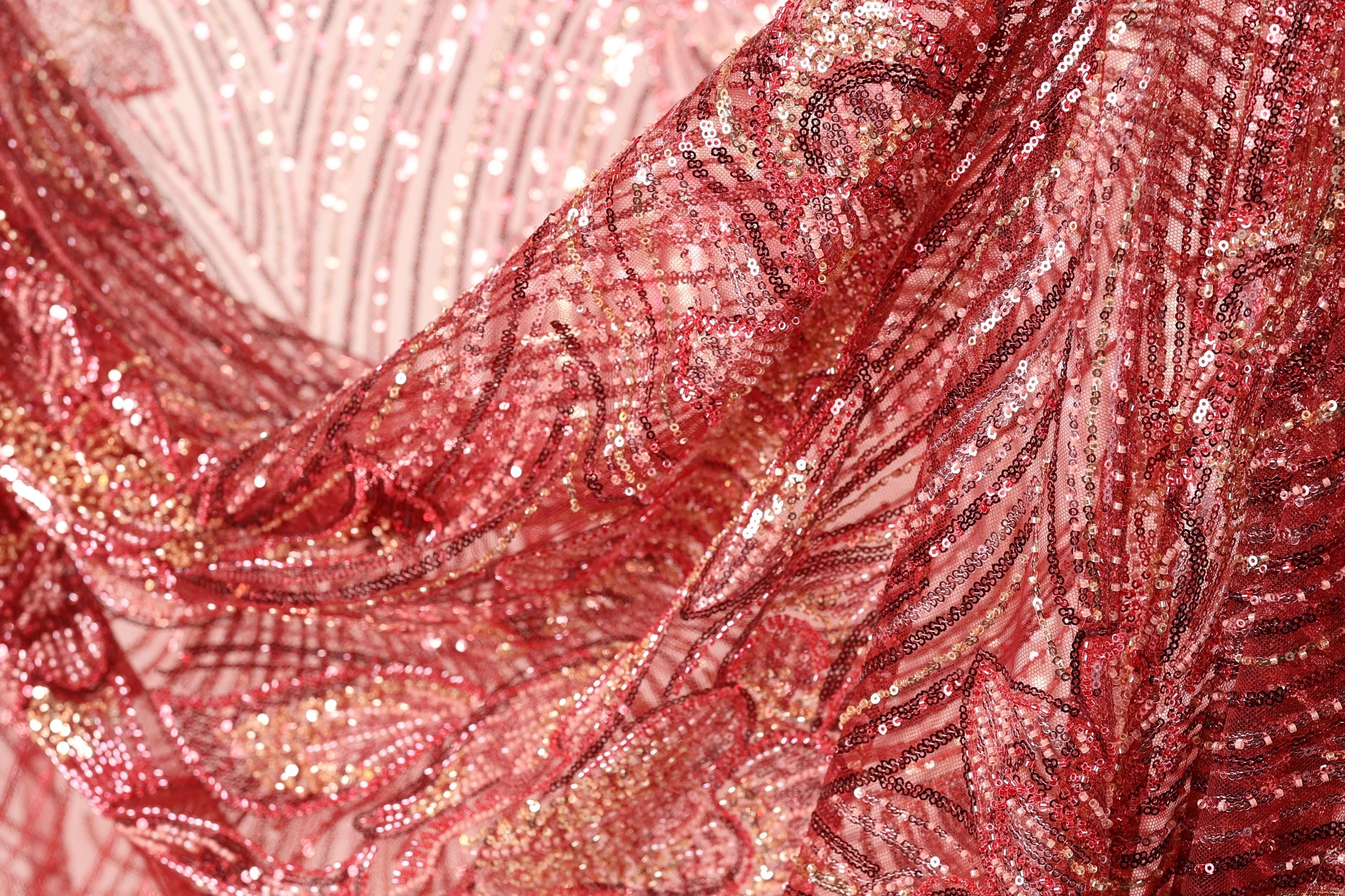 a close-up view of a fabric with a red and pink color scheme, featuring a pattern of sequins and beads. The fabric appears to be made of a shiny, metallic material, giving it a sparkling and luxurious appearance.