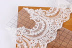 a close-up view of a delicate, intricate lace fabric with a floral pattern. The lace appears to be made of a fine, white thread and is displayed on a wooden surface. The background is blurred, drawing attention to the lace fabric and its details.
