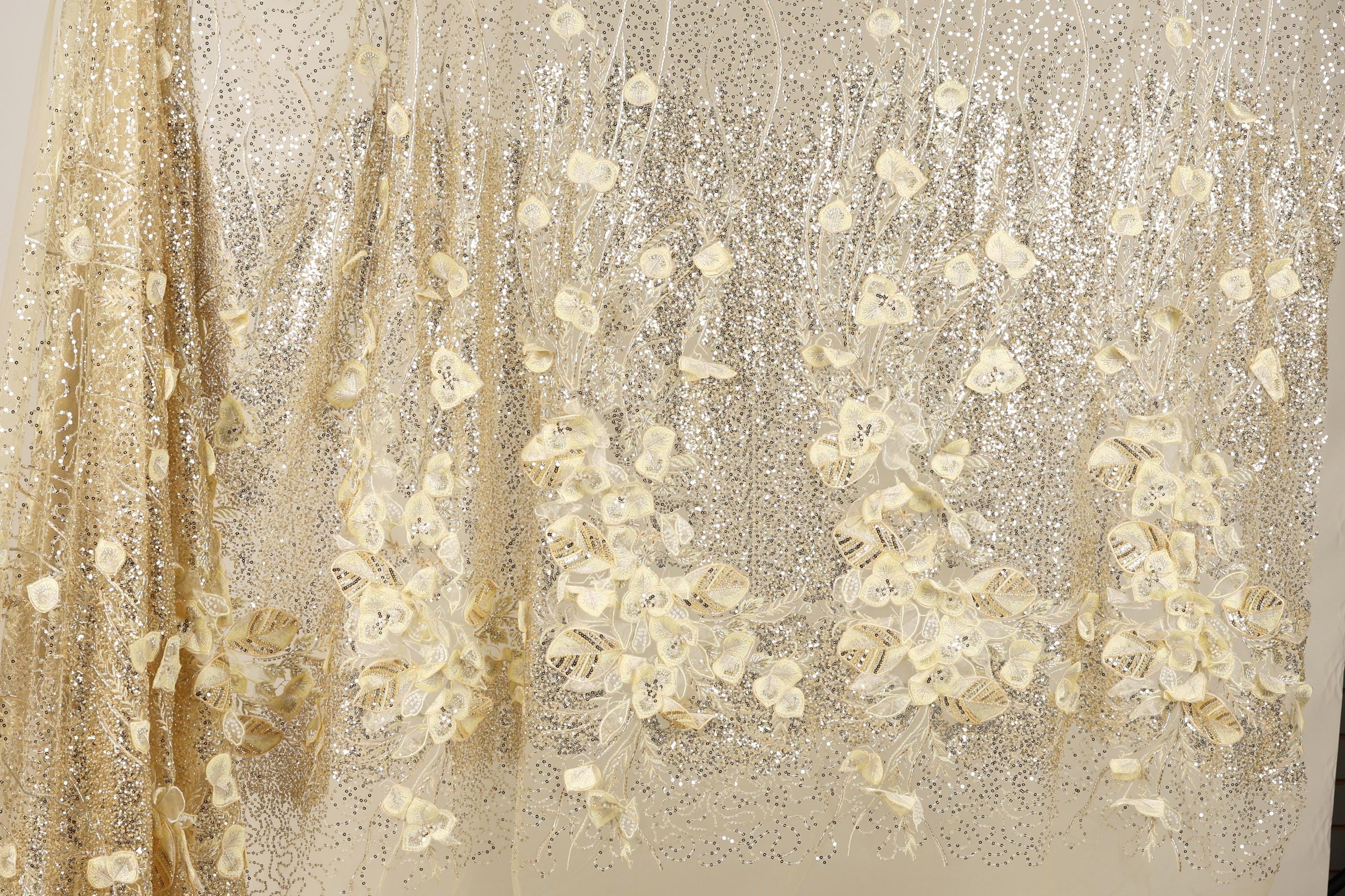 a close-up view of a fabric with a floral pattern, including white and gold flowers. The fabric appears to be made of a shiny, glittery material, giving it a sparkling and elegant appearance.