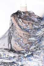 a close-up view of a fabric with a floral pattern, featuring a mix of blue, black, and silver sequins or glitter. The fabric appears to be a sheer or sheer-like material, with a delicate and intricate design.