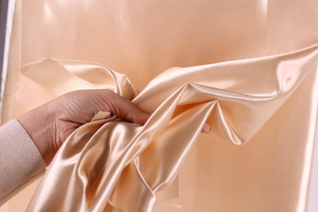 A hand is holding a piece of light brown satin fabric, which appears to be a soft, flowing material.