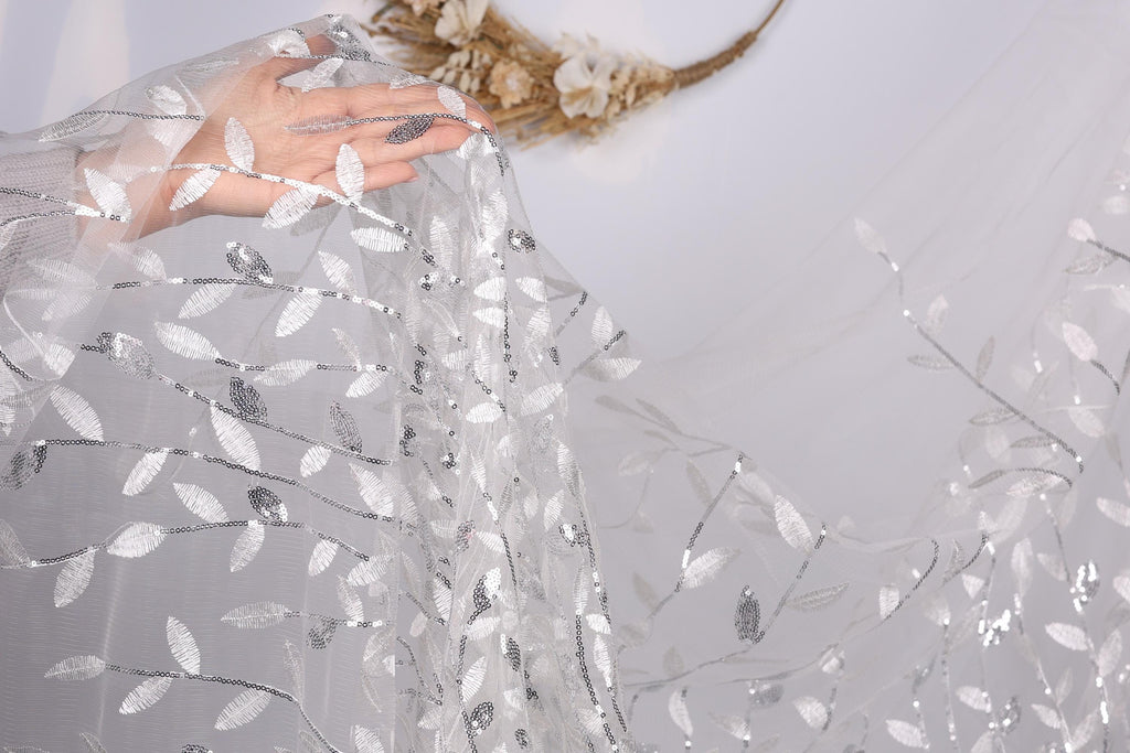 A hand is holding a piece of sheer, white fabric with a leaf pattern, which appears to be a veil or a similar type of garment.