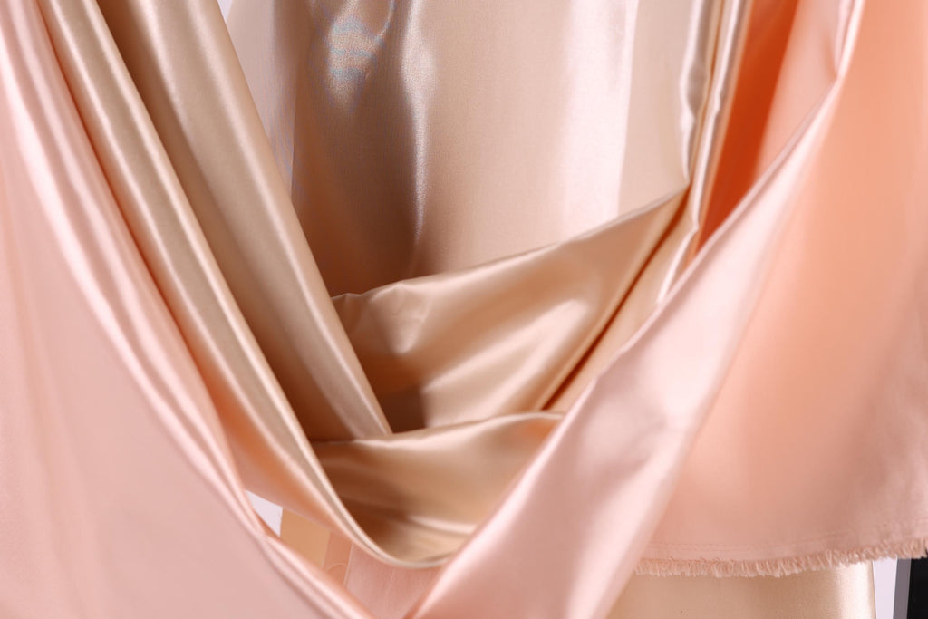 a close-up view of a soft, flowing fabric with a blend of peach and gold colors. The fabric appears to be made of satin or a similar material, with a shiny, smooth texture. The colors are vibrant and the fabric drapes elegantly, creating a visually appealing and luxurious appearance.