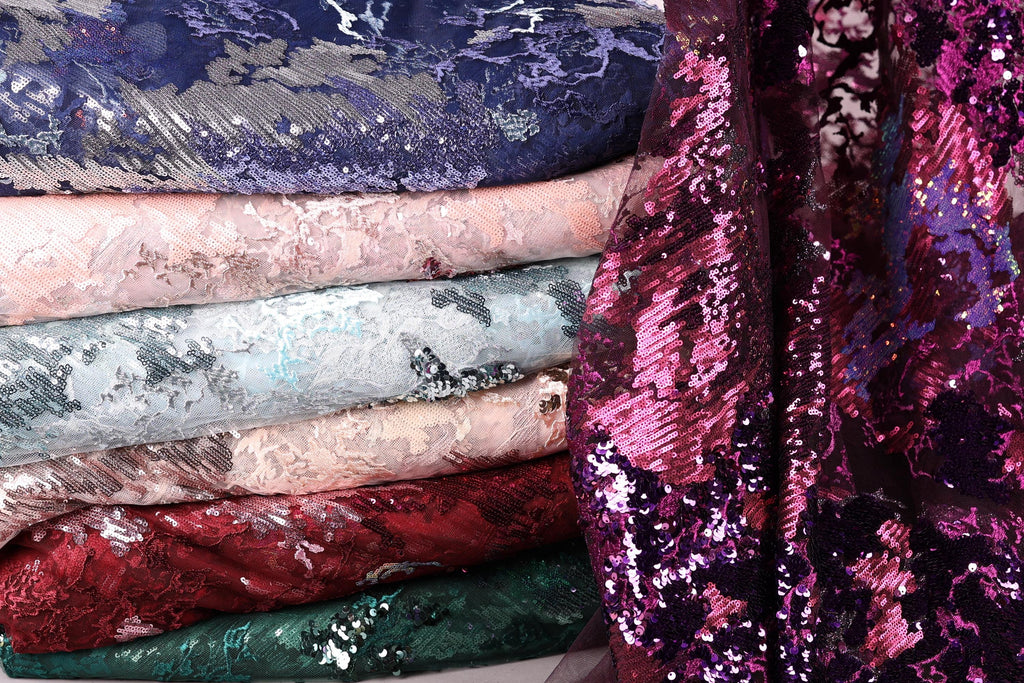 A stack of various colorful fabric swatches, including sequins and lace patterns, with a focus on the vibrant purple and pink hues.