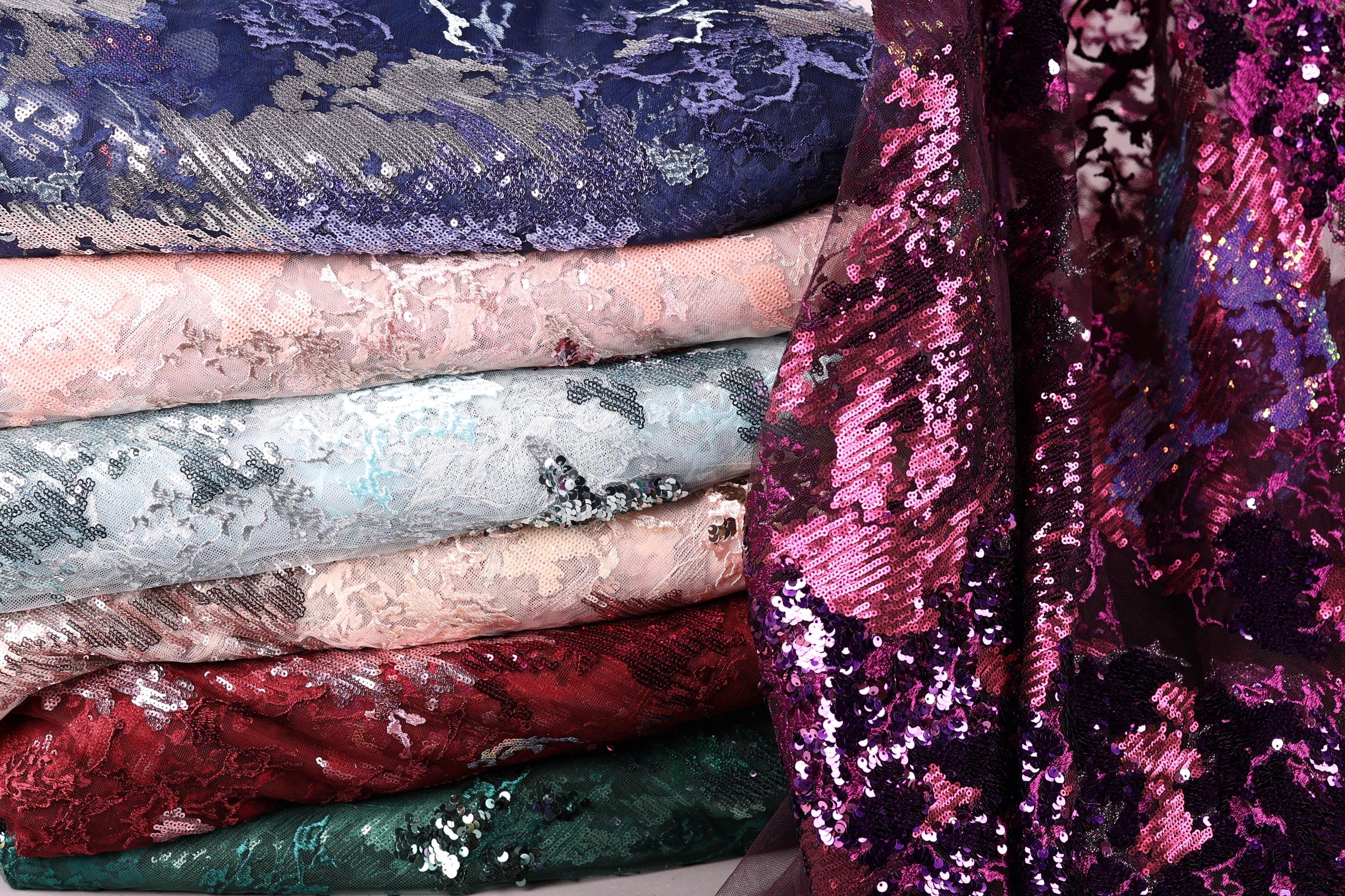 A stack of various colorful fabric swatches, including sequins and lace patterns, with a focus on the vibrant purple and pink hues.