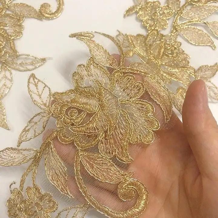 A hand is holding a piece of fabric with intricate gold embroidered floral designs.