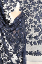 a mannequin wearing a blue lace dress with a floral pattern. The dress is draped over the mannequin, creating a draped effect. The background is a white fabric with a black floral pattern.