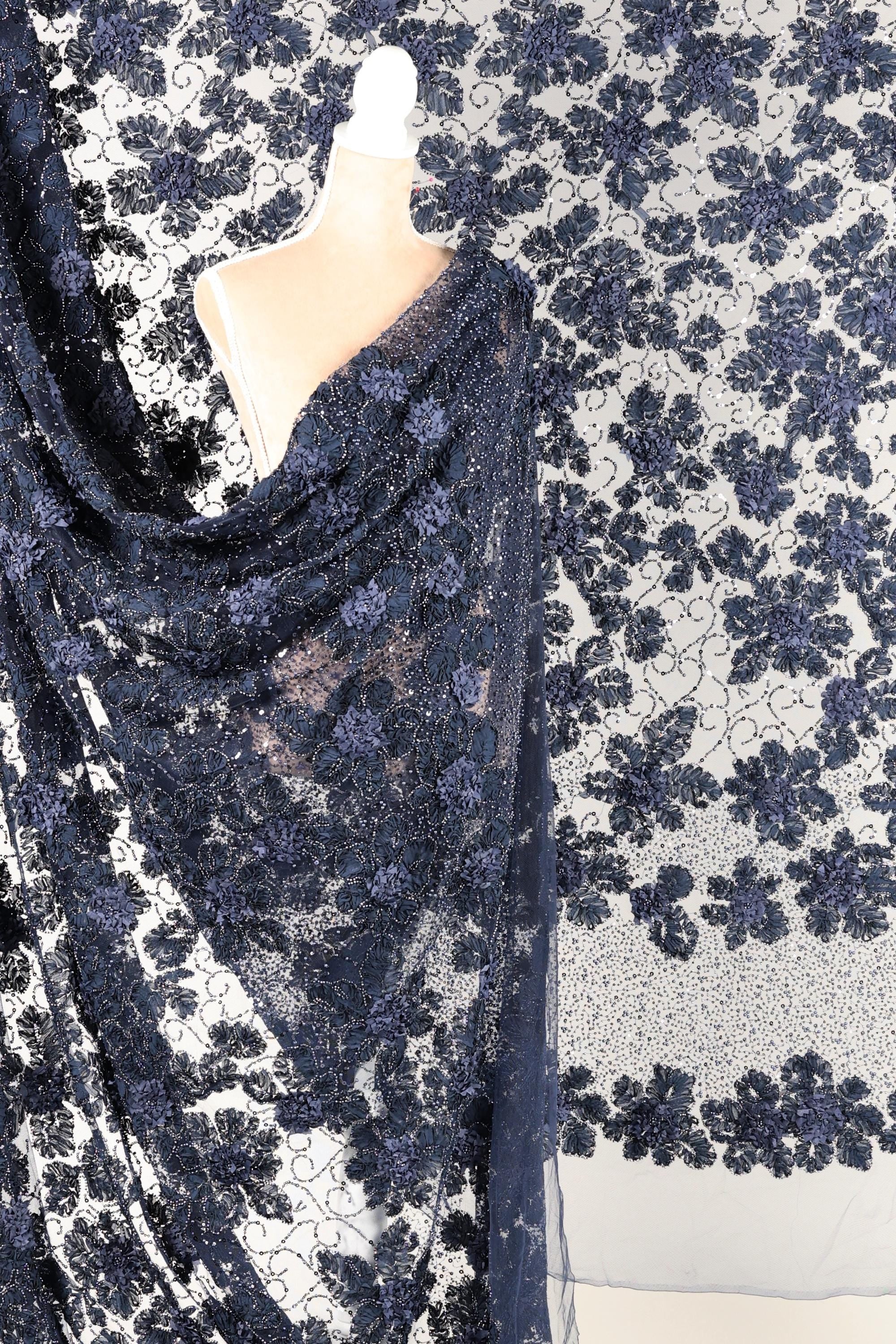 a mannequin wearing a blue lace dress with a floral pattern. The dress is draped over the mannequin, creating a draped effect. The background is a white fabric with a black floral pattern.