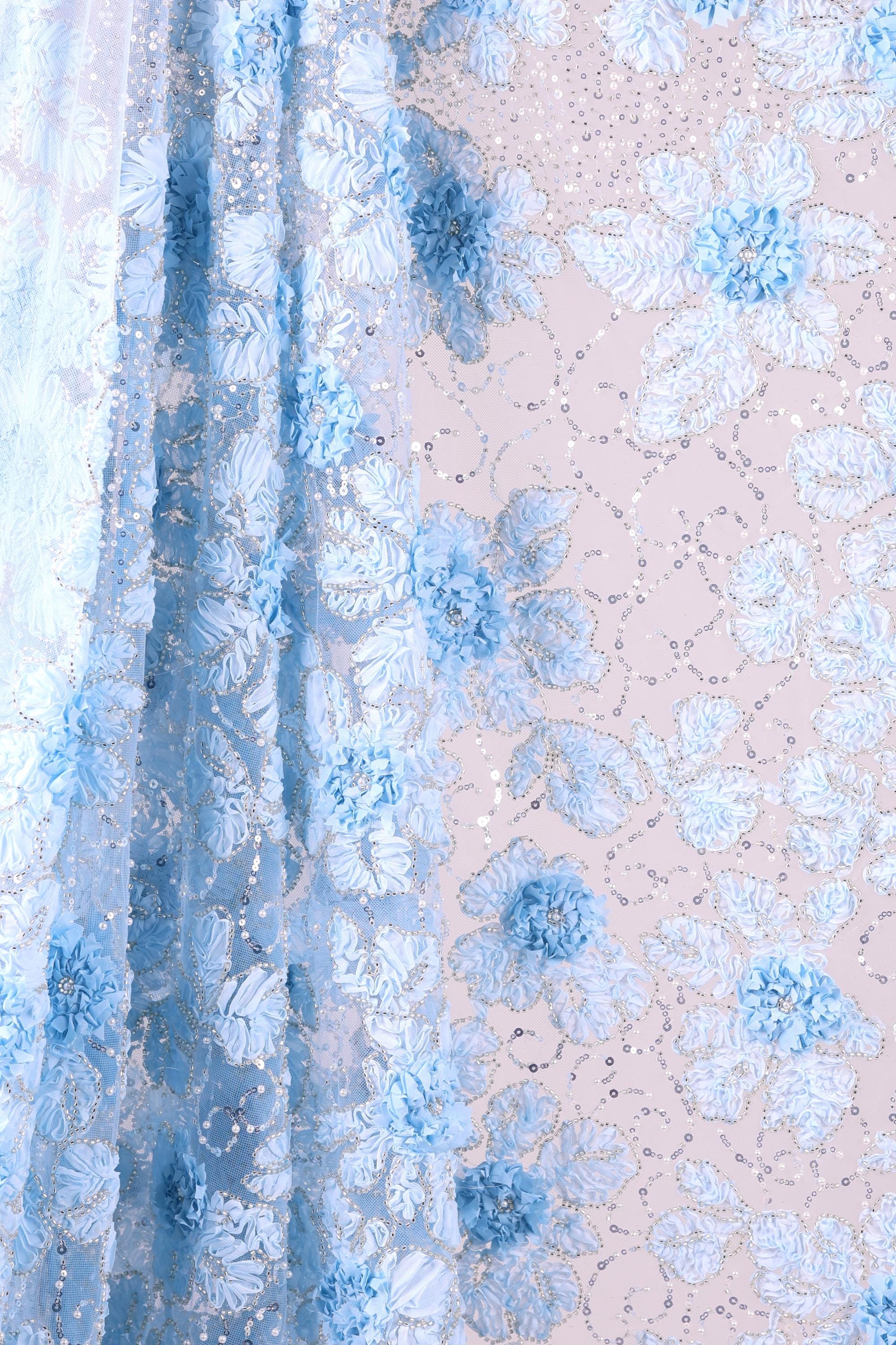 a close-up view of a fabric with a floral pattern in shades of blue and white. The fabric appears to be made of sheer material, with a delicate and intricate design. The pattern consists of various blue flowers and leaves, interspersed with small white dots or sequins, creating a visually appealing and textured appearance.