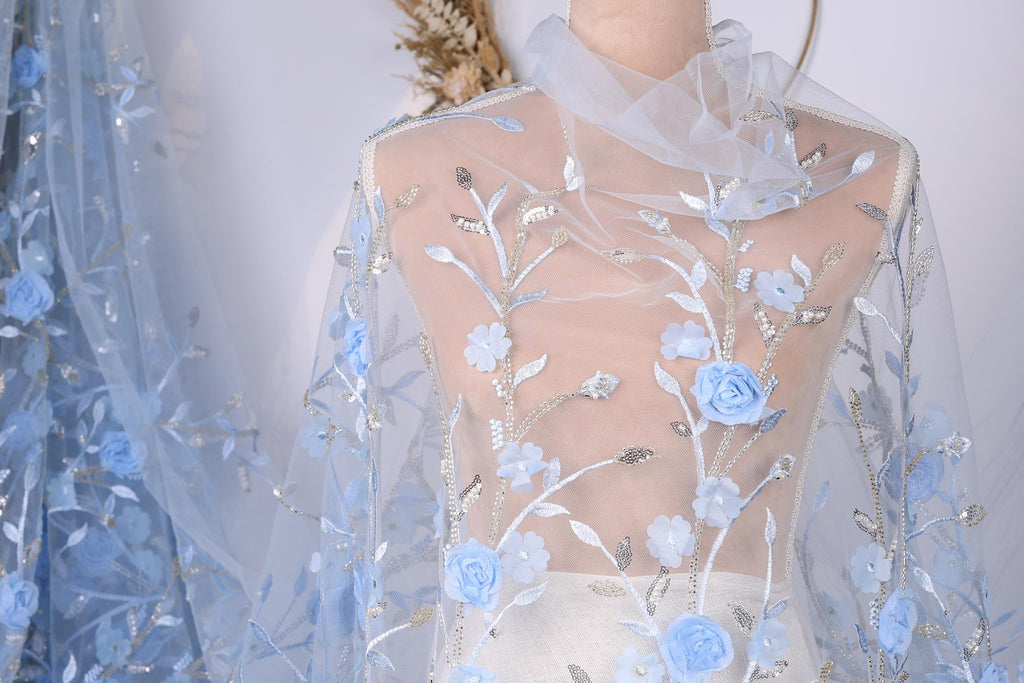 a mannequin wearing a sheer, blue-colored fabric with delicate floral embroidery. The fabric has a light blue color and appears to be made of sheer material, giving it a delicate and airy appearance.