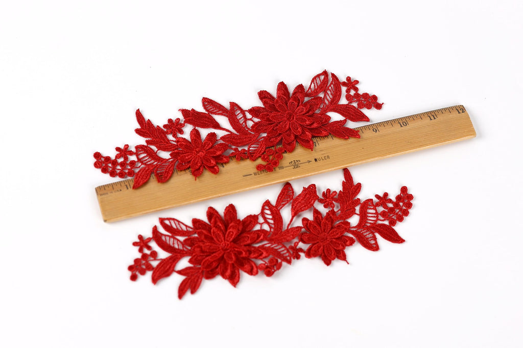 a pair of red floral embroidery pieces lying on a wooden ruler, which is placed on a white background.