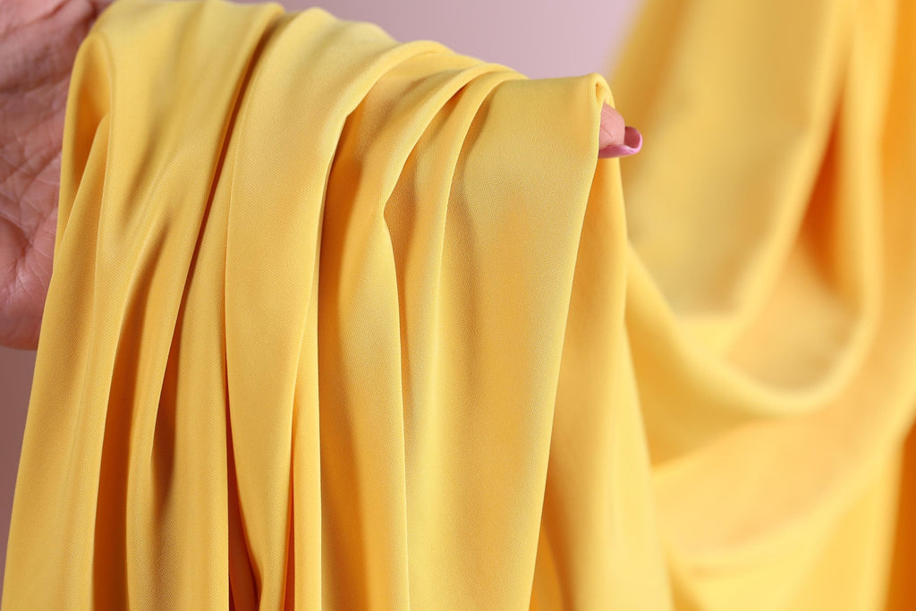 A hand is holding a yellow fabric, which appears to be a piece of clothing or a textile. The fabric is folded and draped, creating a sense of movement and texture.