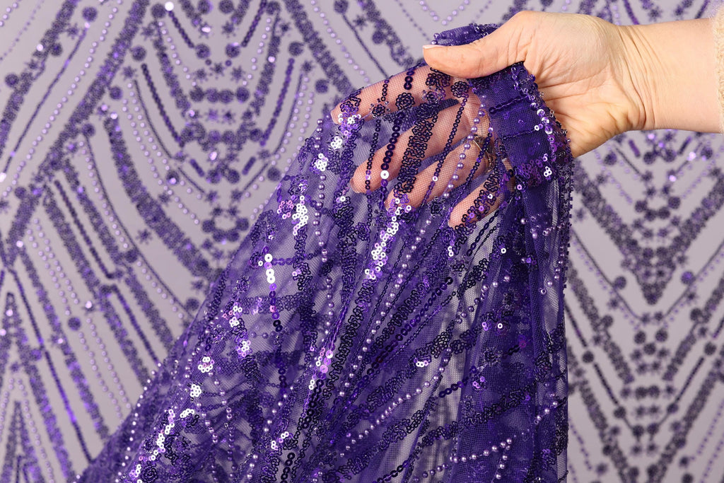 A hand is holding a piece of purple sequin fabric, with the fabric's intricate pattern visible.