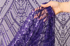 A hand is holding a piece of purple sequin fabric, with the fabric's intricate pattern visible.