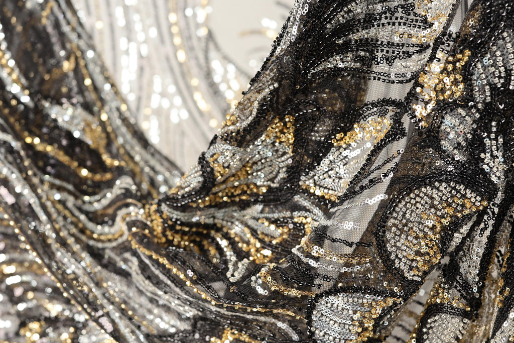 The image is a close-up of a fabric with a black and gold sequin pattern, featuring a mix of black, white, and gold sequins.