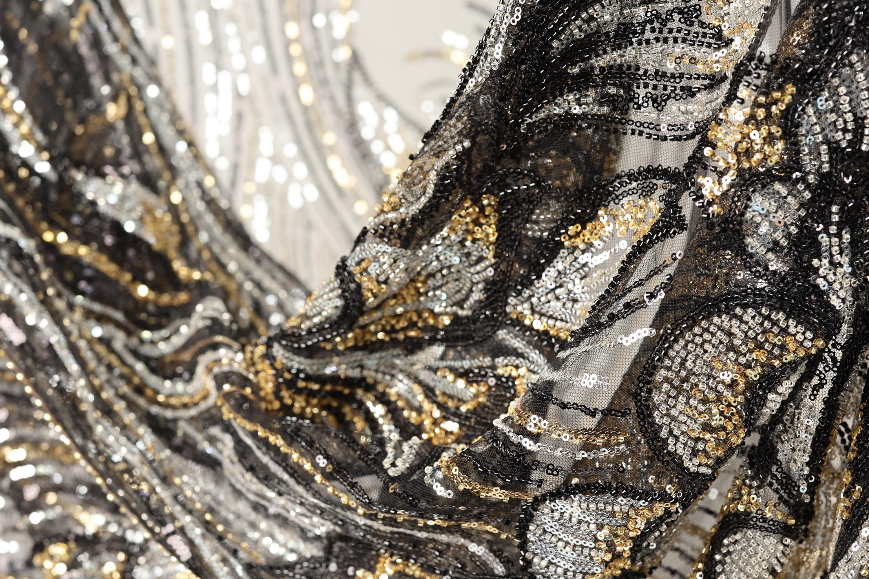 The image is a close-up of a fabric with a black and gold sequin pattern, featuring a mix of black, white, and gold sequins.
