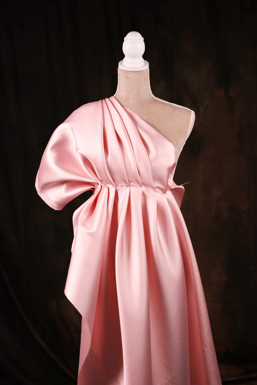 A pink satin one-shoulder dress displayed on a mannequin against a dark background.