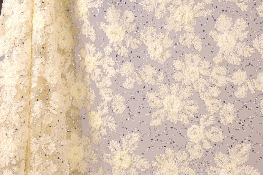 a close-up view of a fabric with a floral pattern, including white and yellow flowers and small silver sequins. The fabric appears to be a sheer material, with a delicate and intricate design.