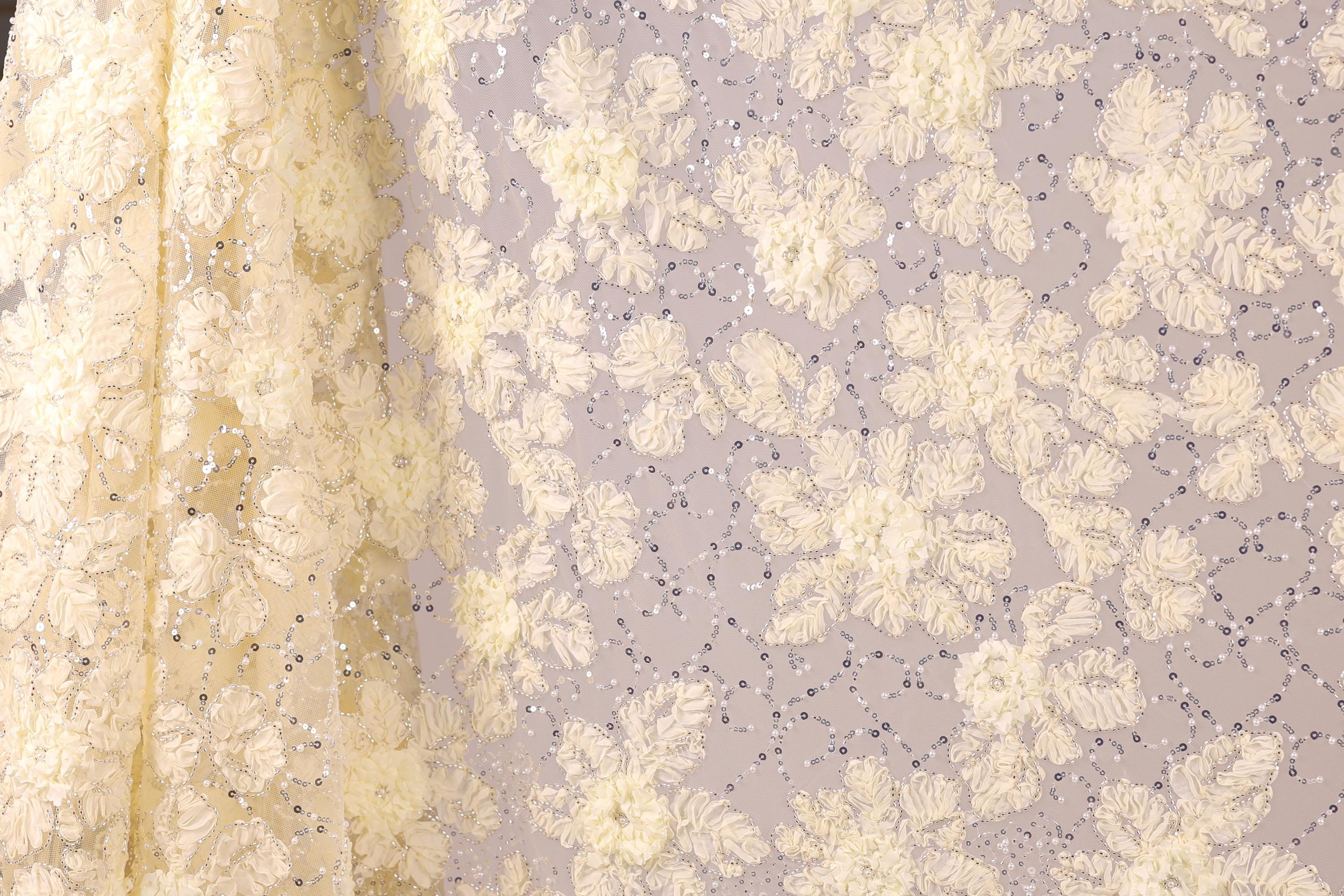 a close-up view of a fabric with a floral pattern, including white and yellow flowers and small silver sequins. The fabric appears to be a sheer material, with a delicate and intricate design.