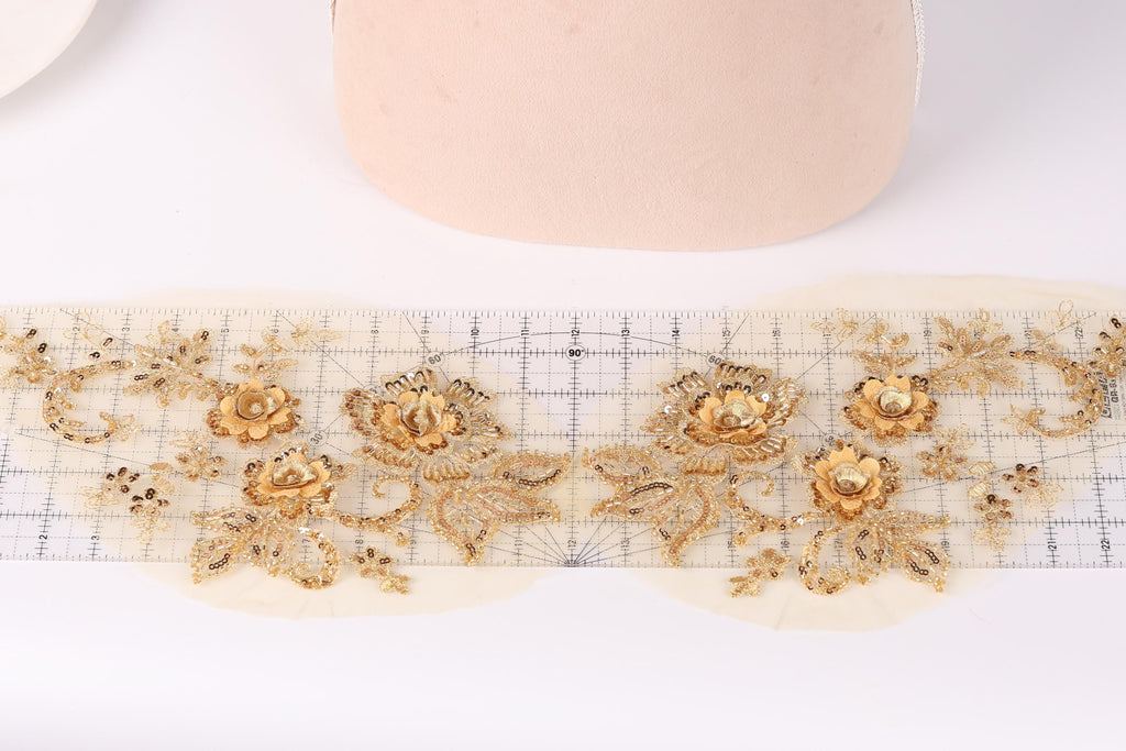 a close-up view of a delicate gold embroidered design on a white background. The design consists of intricate floral patterns and leaves, creating a visually appealing and elegant appearance.