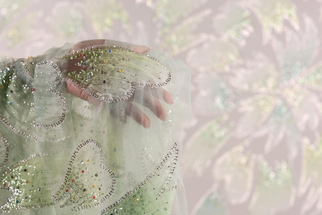 A hand is holding a piece of fabric with a green and silver pattern, against a blurred background.
