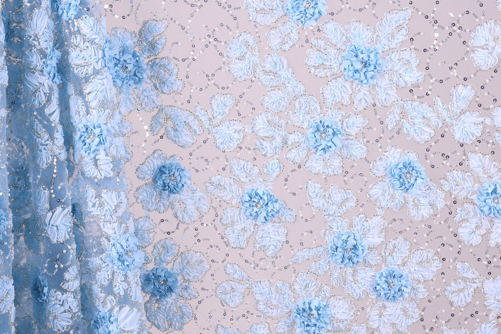 a close-up view of a fabric with a floral pattern, predominantly in shades of blue and white. The fabric appears to be made of a sheer material, with delicate blue flowers and leaves scattered across the surface. The background is a light beige color, providing a neutral backdrop that allows the blue and white floral pattern to stand out.